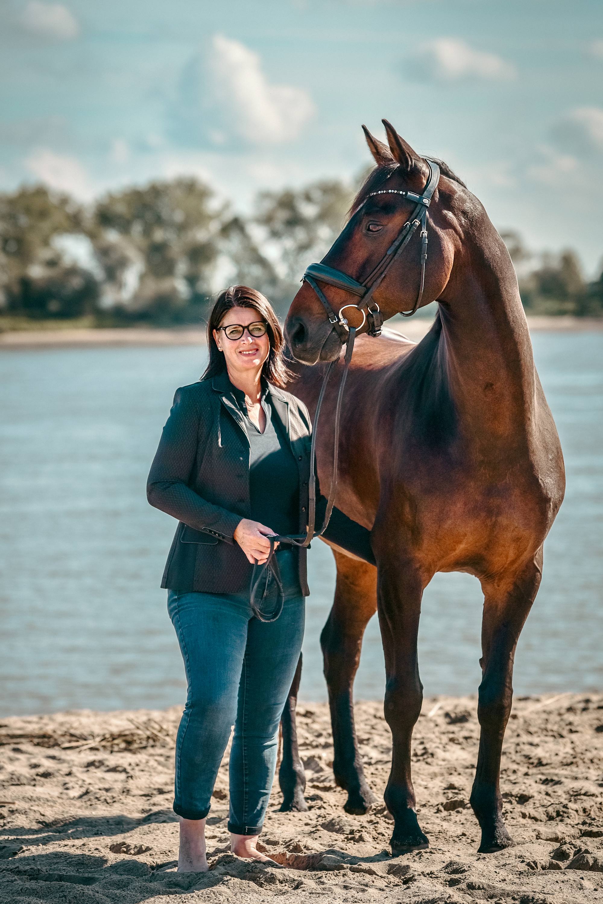 Katja Stöver steht barfuß im flachen Wasser neben ihrem Pferd, im Hintergrund ein weites Gewässer.